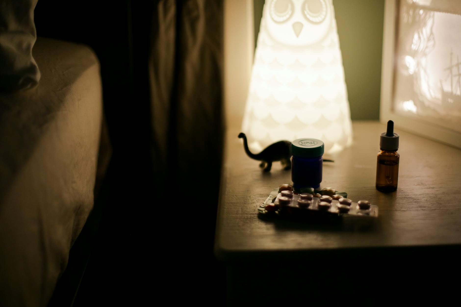 Dimly lit bedside table featuring an owl lamp, medicine bottles, and pill packets, creating a cozy night ambiance.