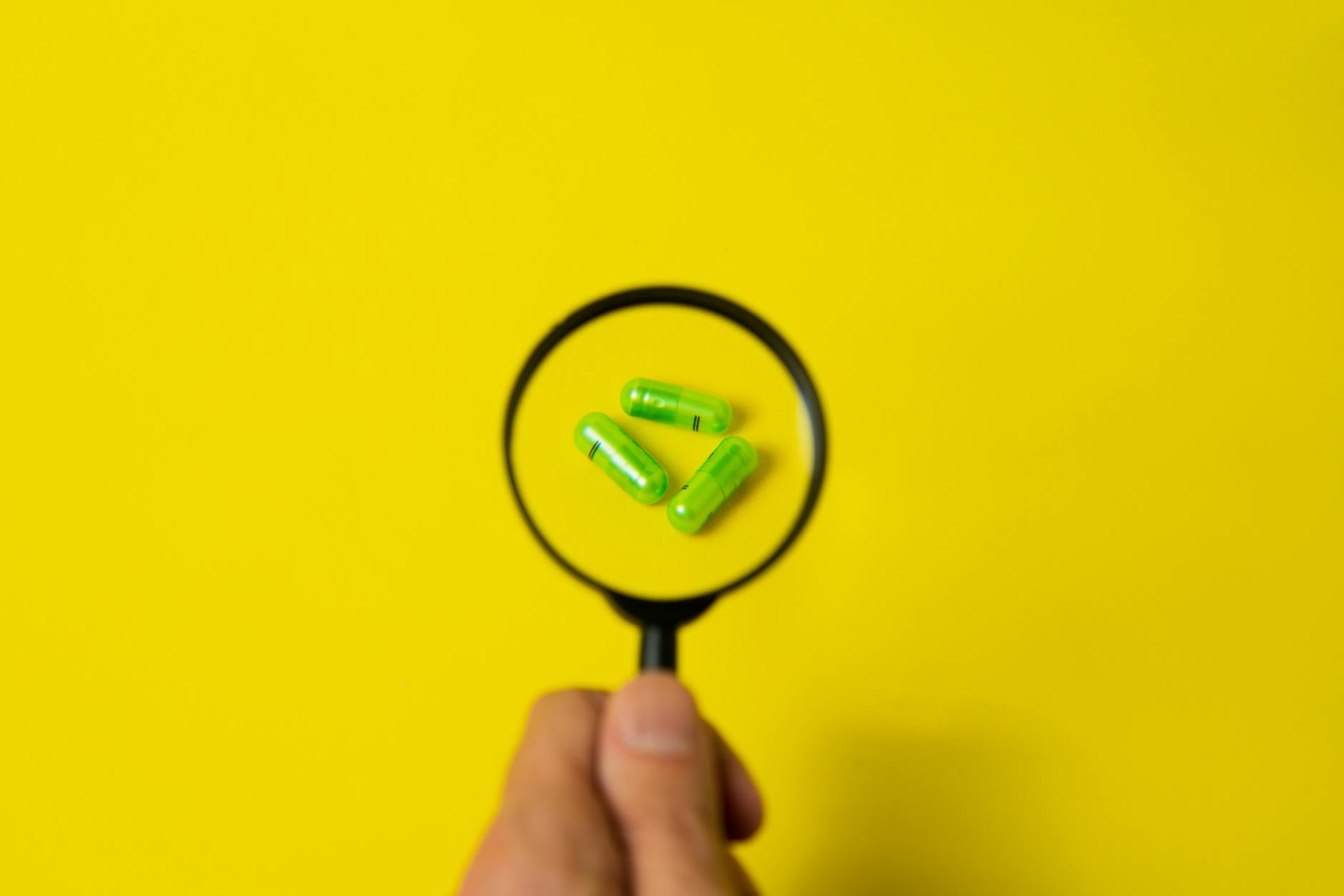 Top view of crop unrecognizable person holding magnifier over green pills placed on bright yellow surface in studio